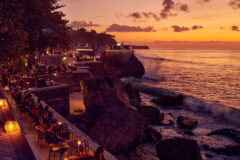 Sunset view of a cliffside restaurant with outdoor seating and people dining along stone terraces overlooking the sea.