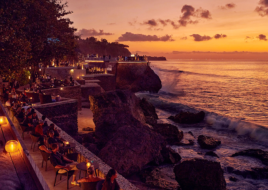Sunset view of a cliffside restaurant with outdoor seating and people dining along stone terraces overlooking the sea.