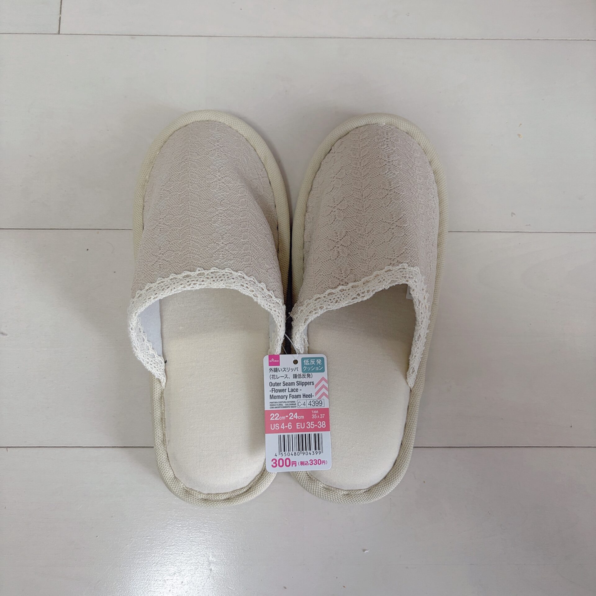 Top-down view of two beige lace-trimmed slippers with a price tag attached on a light wooden floor.