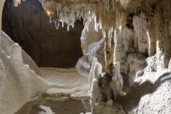 Inside a limestone cave, stalactites hang from the ceiling while stalagmites rise from the sandy floor, forming a pale cavern.