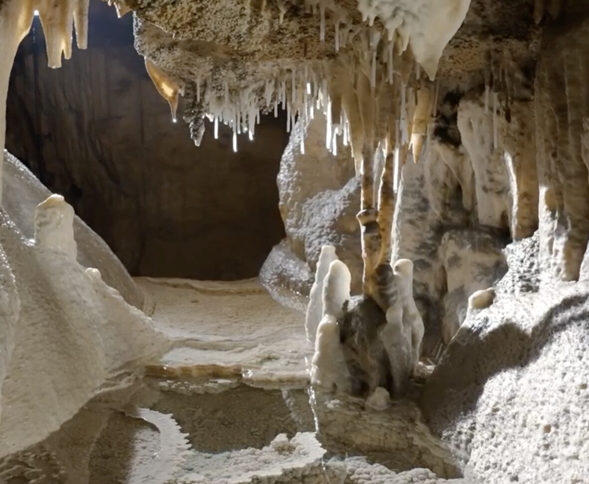 Inside a limestone cave, stalactites hang from the ceiling while stalagmites rise from the sandy floor, forming a pale cavern.