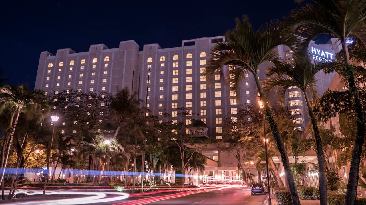 Night view of the Hyatt Regency hotel at night, with illuminated windows, palm trees, and light trails from passing cars in the foreground.