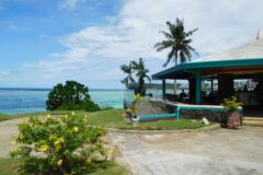 Open-air seaside restaurant with turquoise roof and glass railing overlooking clear blue water and palm trees on a sunny day (tropical setting).