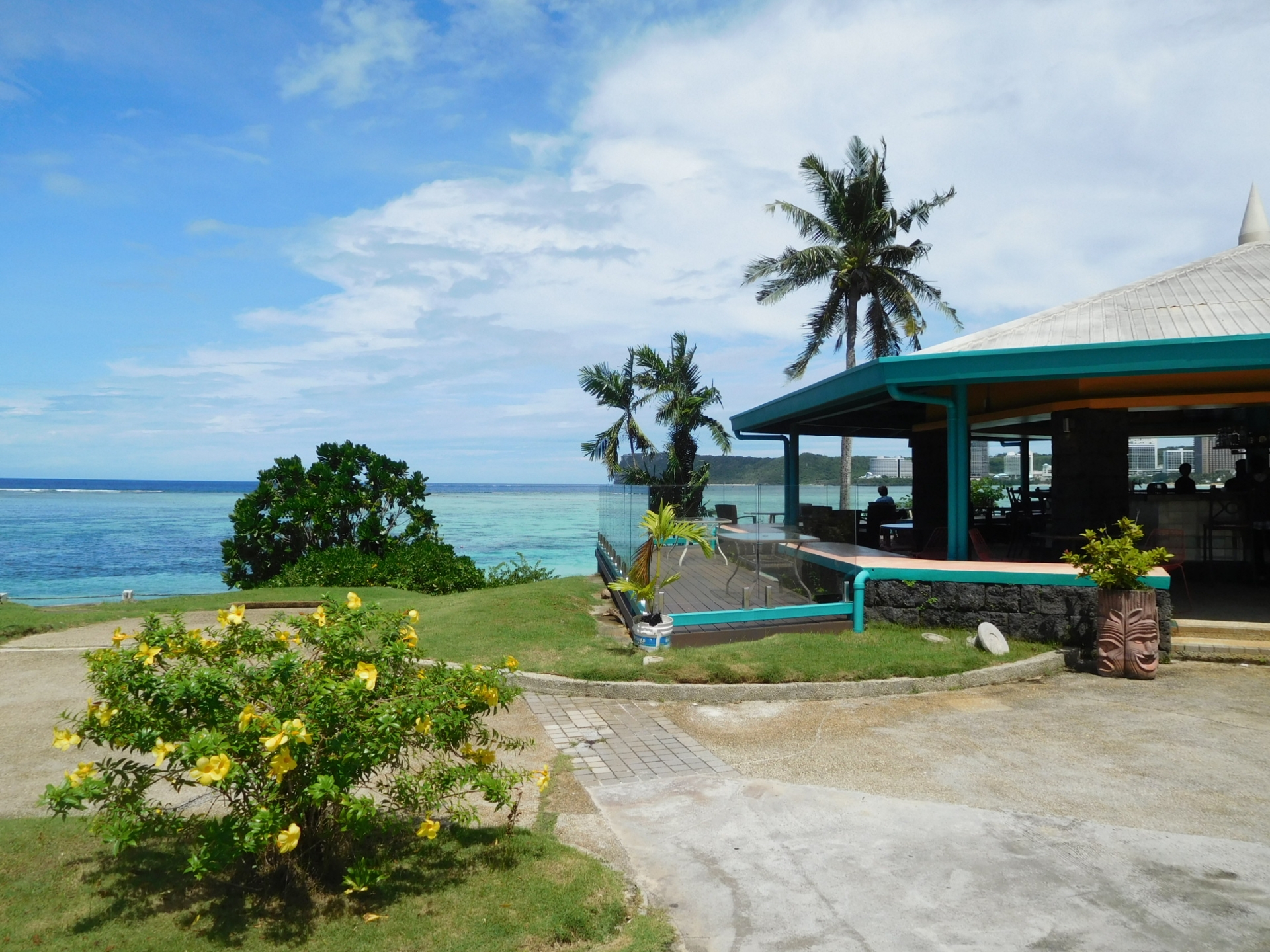 Open-air seaside restaurant with turquoise roof and glass railing overlooking clear blue water and palm trees on a sunny day (tropical setting).
