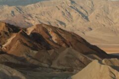 Arid desert landscape with layered brown and tan sand dunes and rugged mountains in the distance.