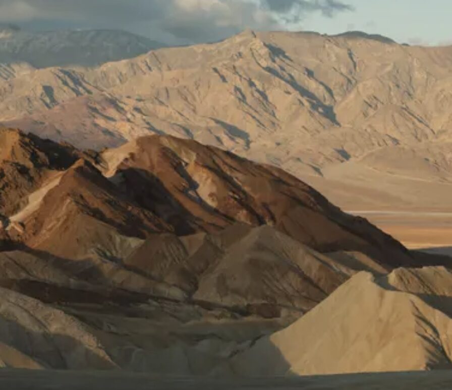 Arid desert landscape with layered brown and tan sand dunes and rugged mountains in the distance.