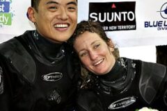 Two divers in black wetsuits smiling with heads together, posing after a dive event with sponsor banners in the background.