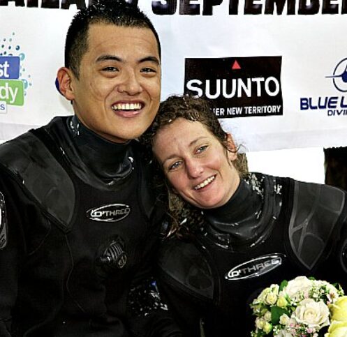 Two divers in black wetsuits smiling with heads together, posing after a dive event with sponsor banners in the background.