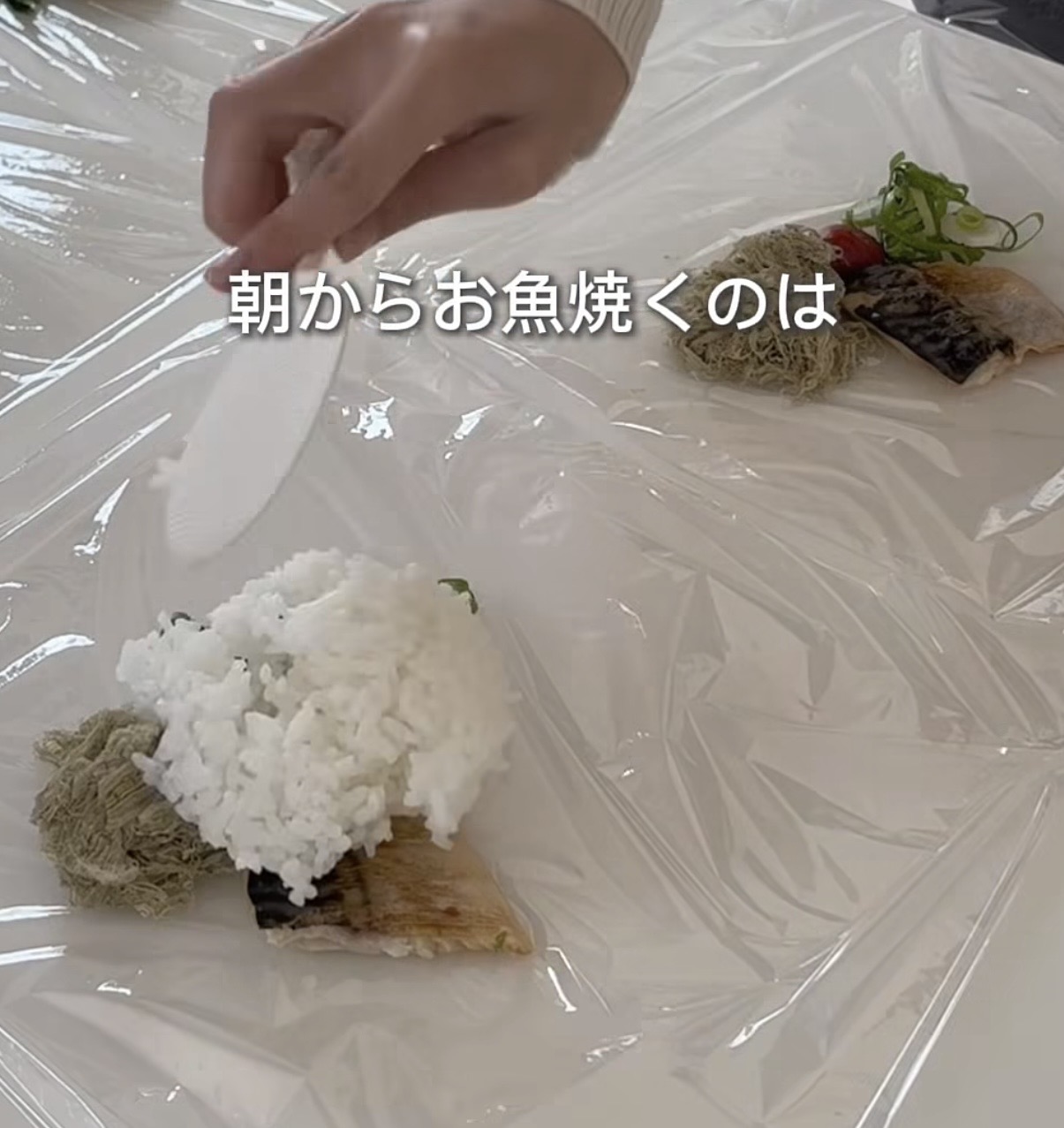 Screenshot | GLAM(グラム)|自分らしく生きる女性に、1日1GLAMを。 A hand holds a spatula over a sushi-style piece with white rice on top placed on plastic wrap, with greens and other toppings nearby; Japanese caption reads'朝からお魚焼くのは'.
