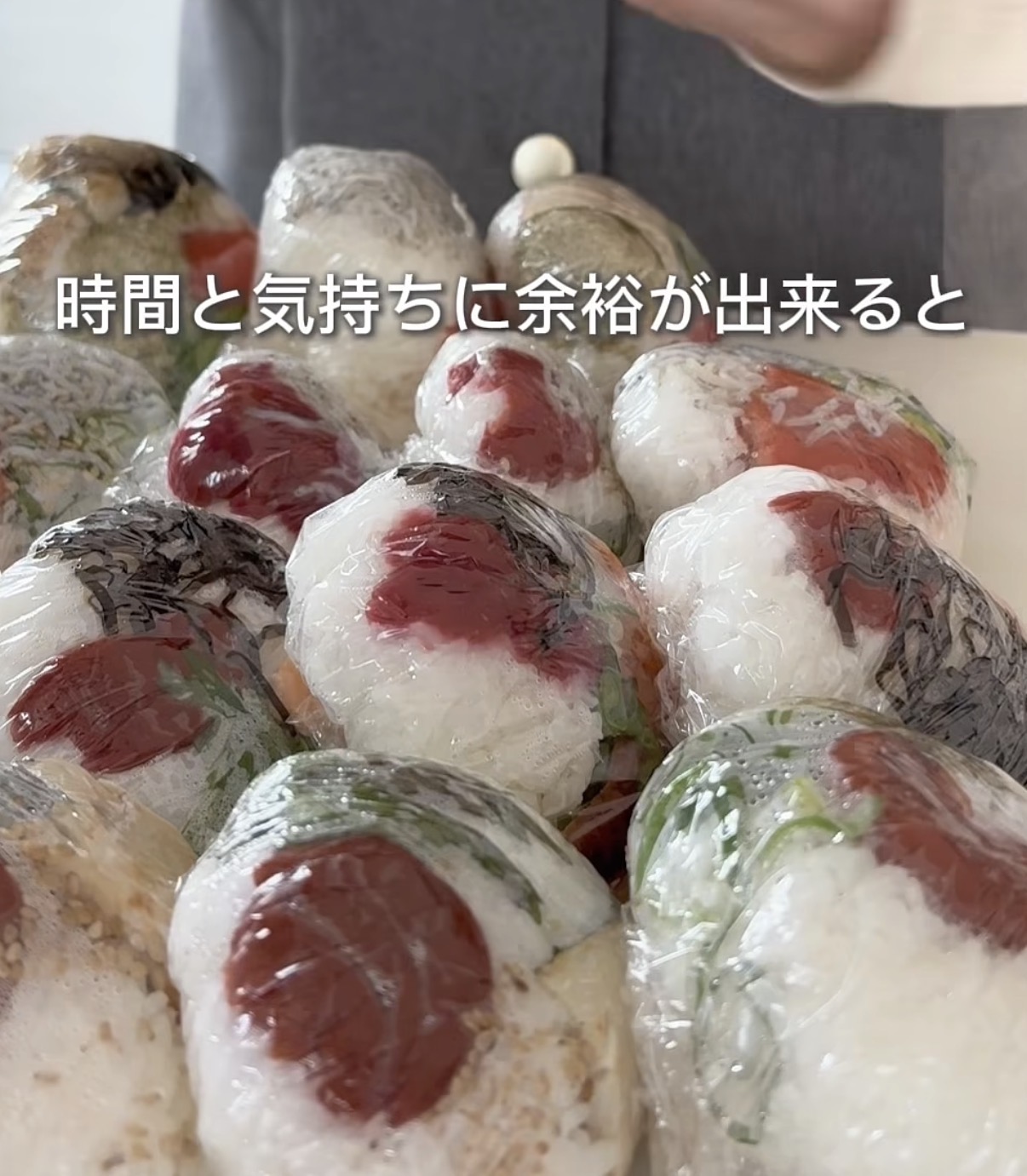 Screenshot | GLAM(グラム)|自分らしく生きる女性に、1日1GLAMを。 Pile of plastic-wrapped onigiri (Japanese rice balls) with red fillings, ready to eat. Text overlay reads Japanese phrase about having time and space.