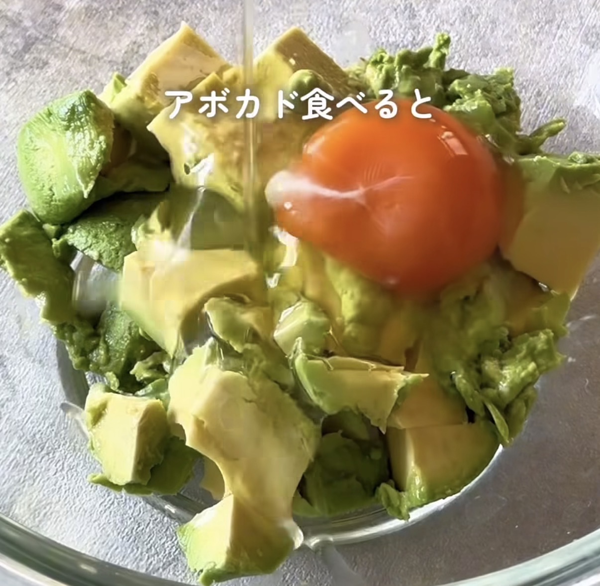 Chopped avocado and greens in a glass bowl with a tomato on top and a drizzle of oil; Japanese caption over the image says 'When you eat avocado'.