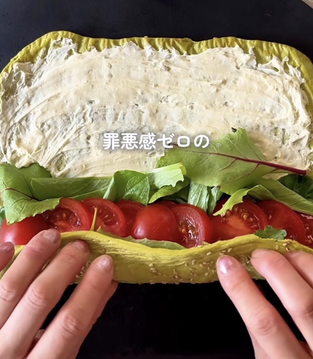 Wrap sandwich being assembled: yellow flatbread filled with cream cheese spread, lettuce, and sliced tomatoes, held open by hands.