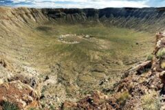 Wide view of a large circular crater with a green grassy floor and rugged rocky rim under a blue sky.