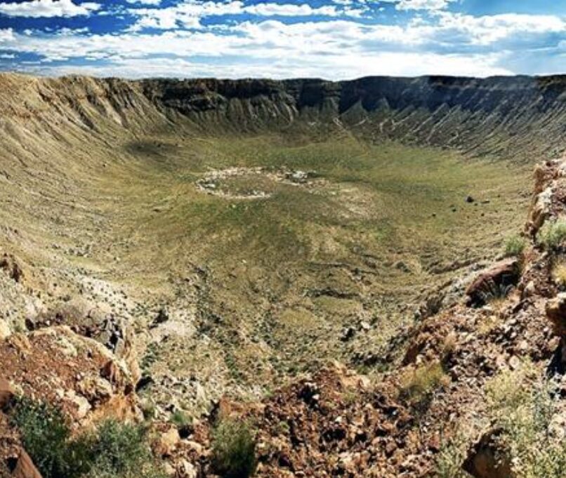 Wide view of a large circular crater with a green grassy floor and rugged rocky rim under a blue sky.