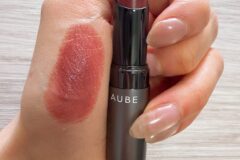 Close-up of a lipstick being applied to lips, with swatched lipstick on the back of the hand and the tube showing the brand name AUBE.