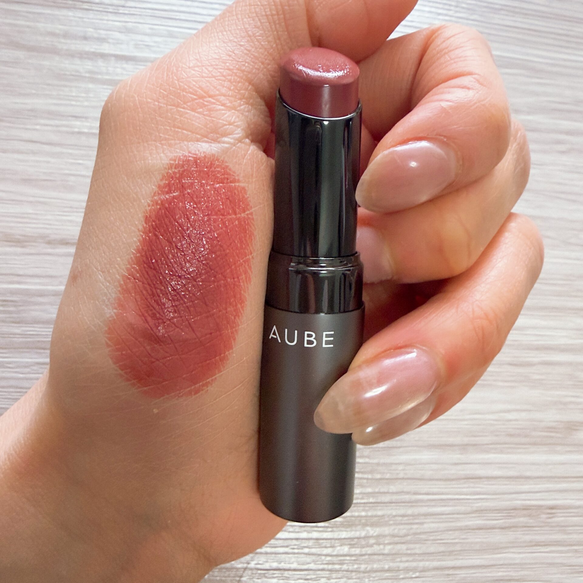 Close-up of a lipstick being applied to lips, with swatched lipstick on the back of the hand and the tube showing the brand name AUBE.