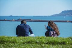 Two people sit on a grassy embankment by a blue sea; the woman holds a smartphone while the man gazes toward the water.