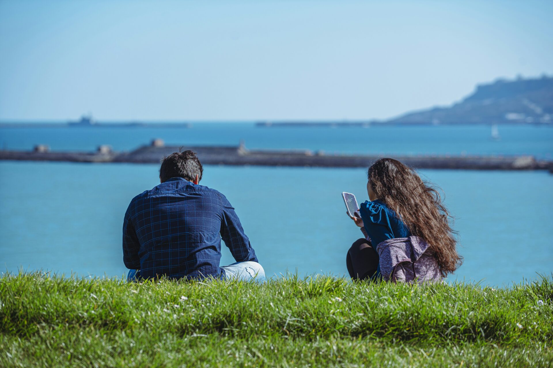 Two people sit on a grassy embankment by a blue sea; the woman holds a smartphone while the man gazes toward the water.