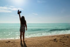 A young girl stands barefoot on a sandy beach, raising a blue hat toward the bright sky as turquoise waves wash in.