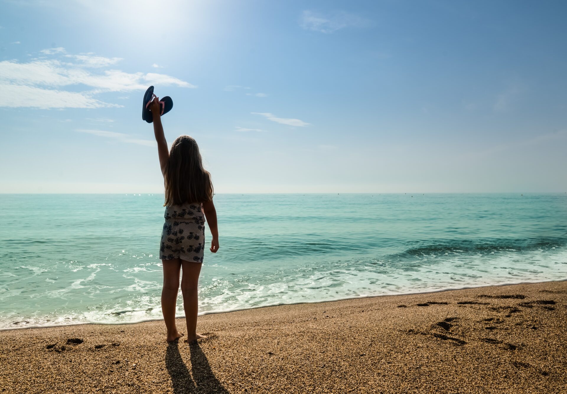 A young girl stands barefoot on a sandy beach, raising a blue hat toward the bright sky as turquoise waves wash in.