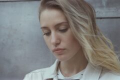 Close-up of a woman with blonde hair looking down, wearing a light jacket against a gray wall background.