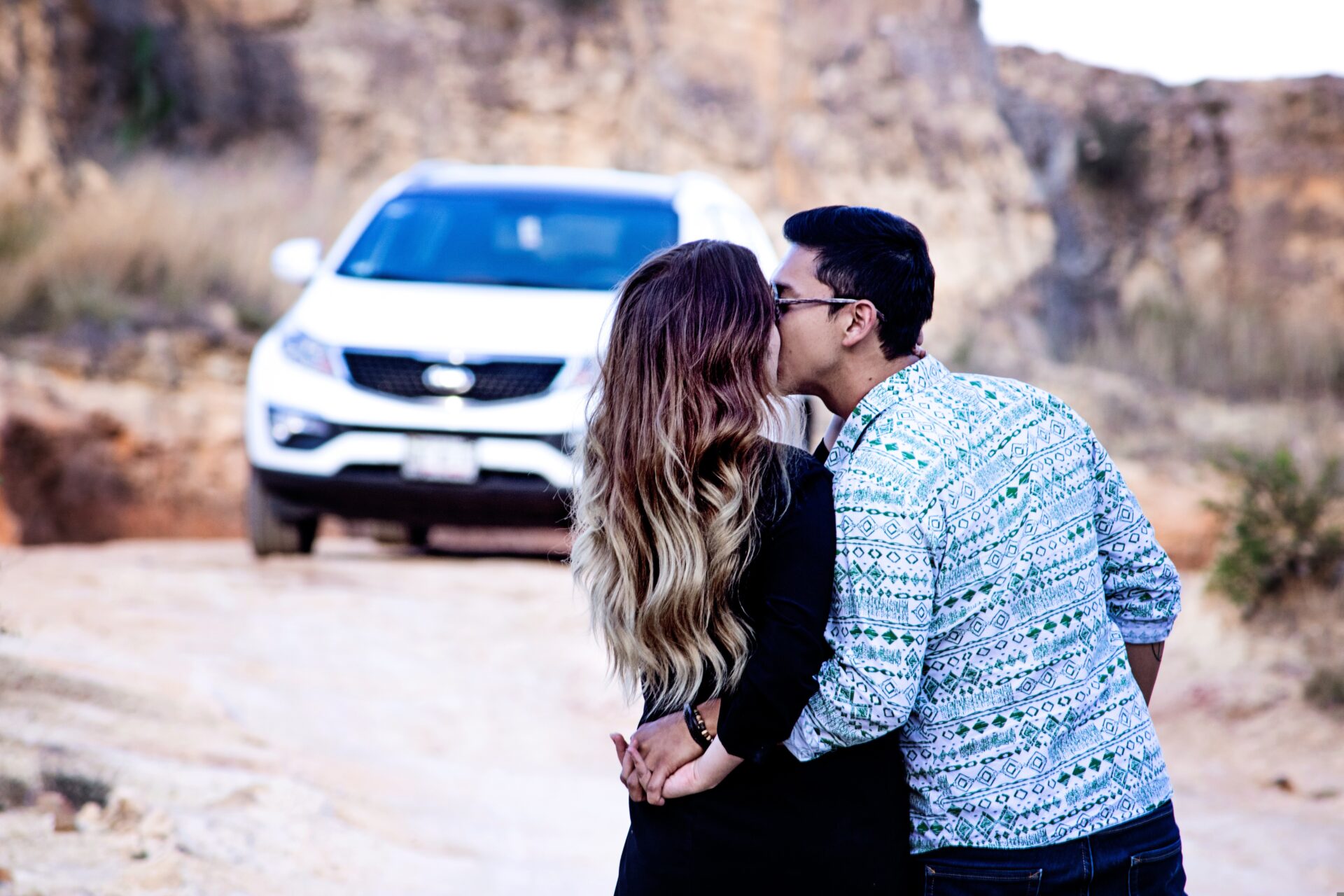Couple kissing in an outdoor rocky area with a white SUV in the background.