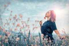 Woman standing in a field of tall pink flowers, holding a red blossom near her lips with a bright sun halo behind her.