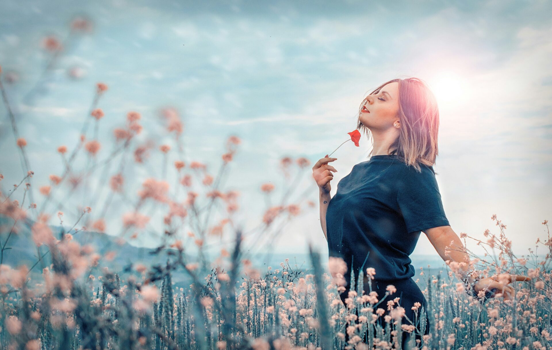 Woman standing in a field of tall pink flowers, holding a red blossom near her lips with a bright sun halo behind her.
