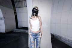 Person wearing a black mask and white tank top standing against a concrete pillar in a gray urban tunnel, ripped jeans shown.