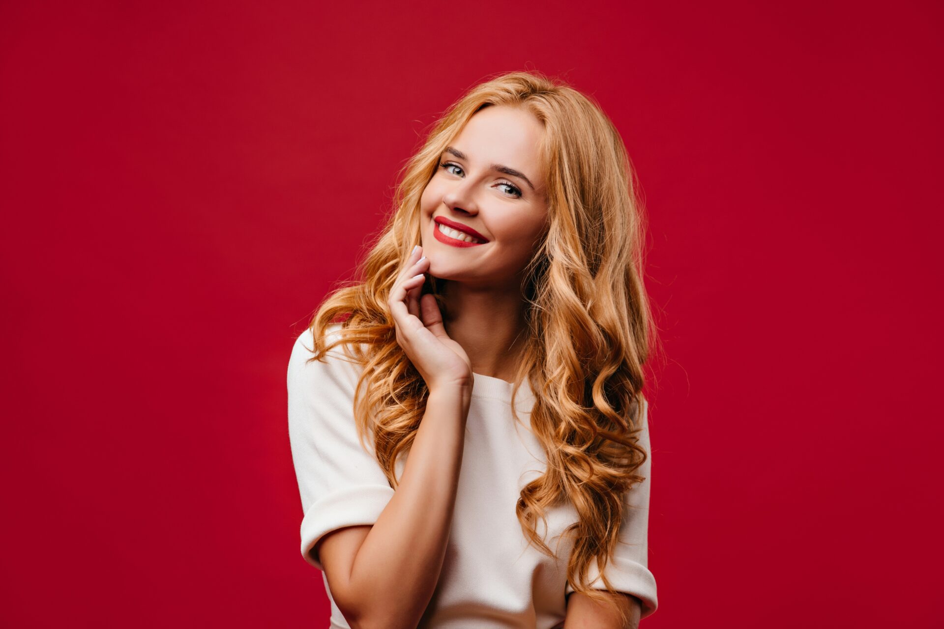 Smiling blonde woman with wavy hair in a white top, posed against a red background.