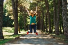 Woman wearing teal shirt and jeans jumps mid-air on a sunlit forest path, arms raised in joy with red sneakers visible behind trees.