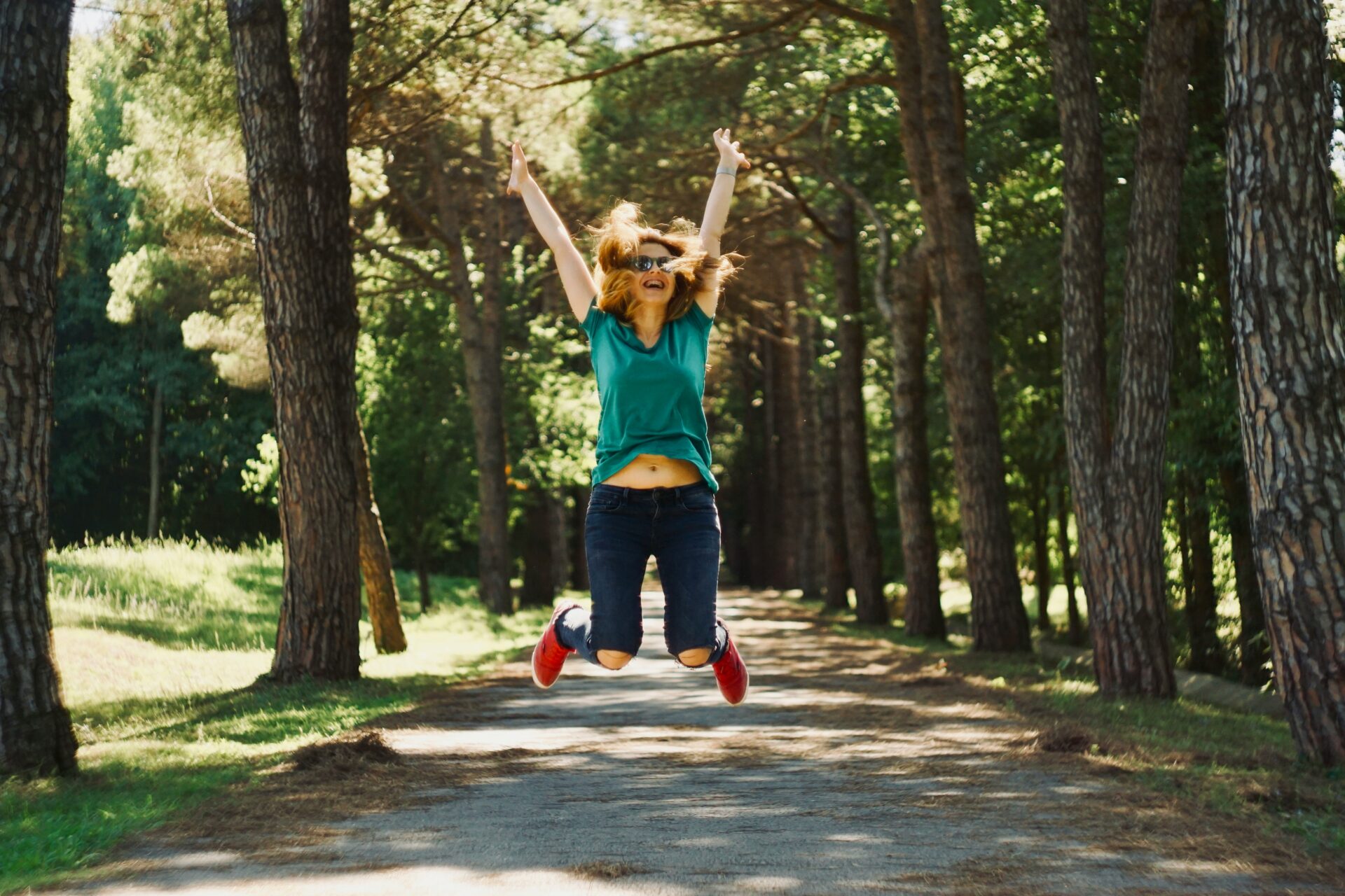 Woman wearing teal shirt and jeans jumps mid-air on a sunlit forest path, arms raised in joy with red sneakers visible behind trees.