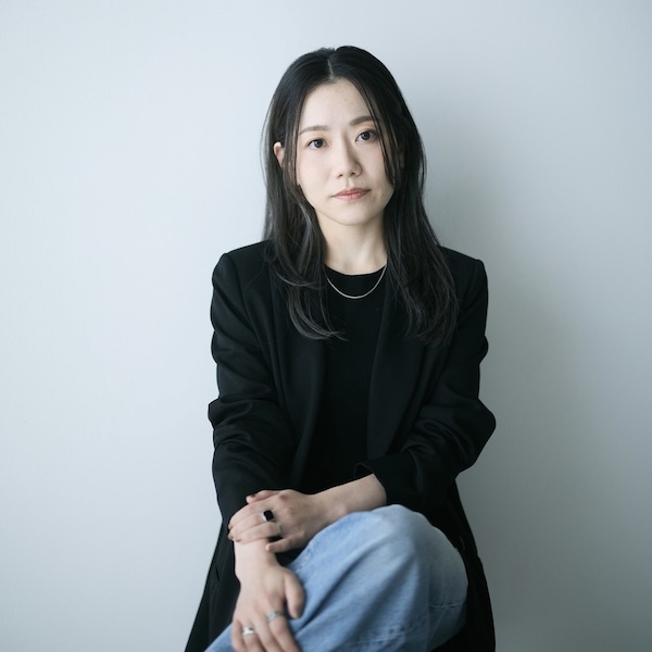 Portrait of a woman with long black hair, wearing a black blazer and light jeans, sitting against a pale wall with a calm expression.