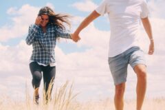 Couple running through tall grass, holding hands under a bright blue sky with clouds.