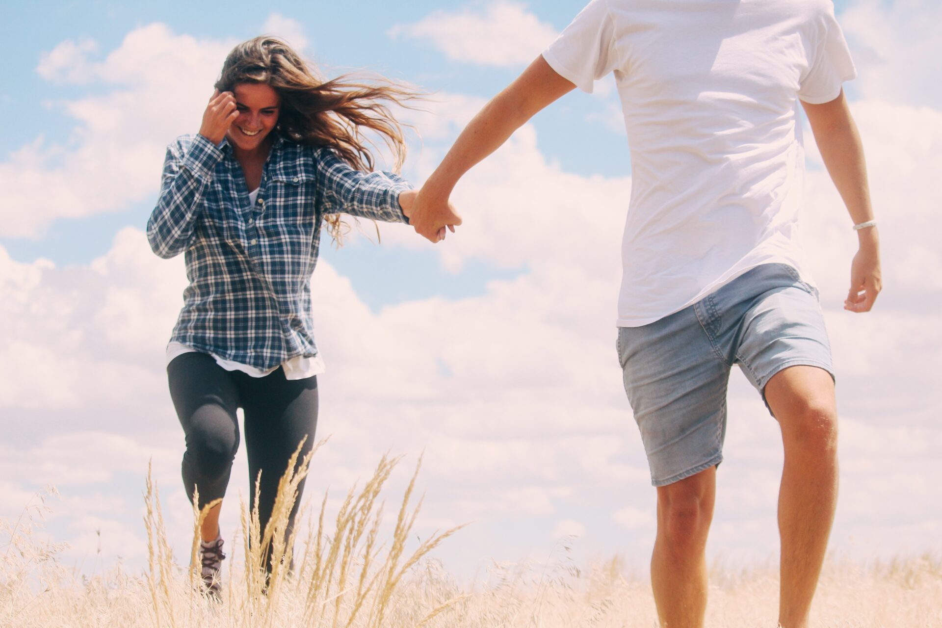 Couple running through tall grass, holding hands under a bright blue sky with clouds.