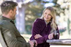 Woman with blonde hair in a purple fleece smiles at a man while holding a steaming mug at a park picnic table.