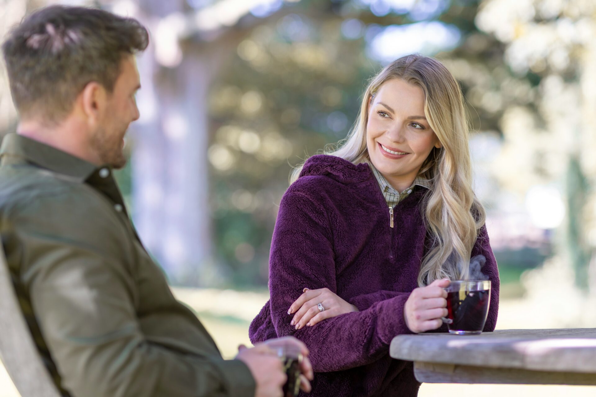 Woman with blonde hair in a purple fleece smiles at a man while holding a steaming mug at a park picnic table.