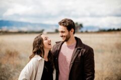 Couple laughing and looking at each other in a sunlit field with mountains in the background.
