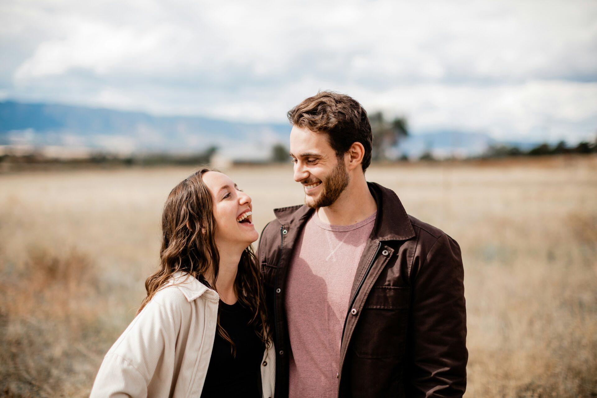 Couple laughing and looking at each other in a sunlit field with mountains in the background.