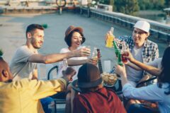 Group of friends on a rooftop toasting drinks around a table with chips and a guitar in the background, sunset vibe.