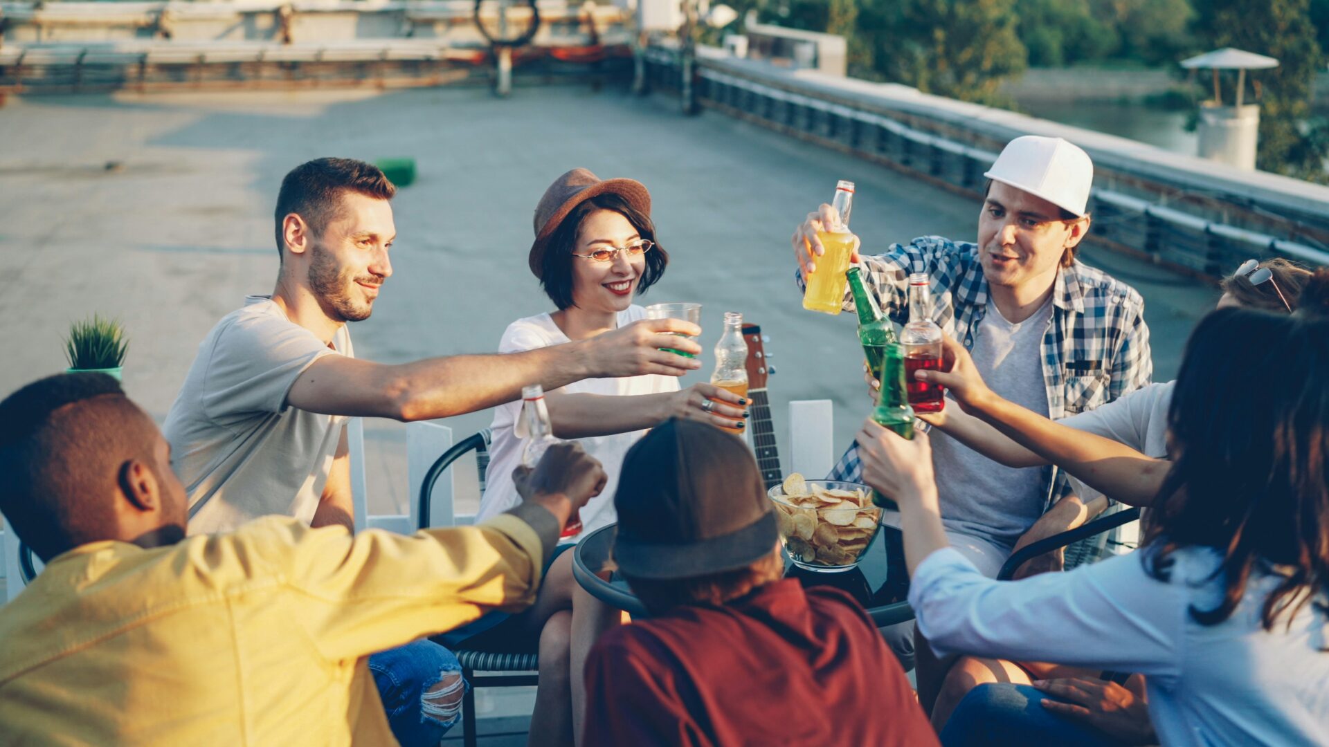 Group of friends on a rooftop toasting drinks around a table with chips and a guitar in the background, sunset vibe.
